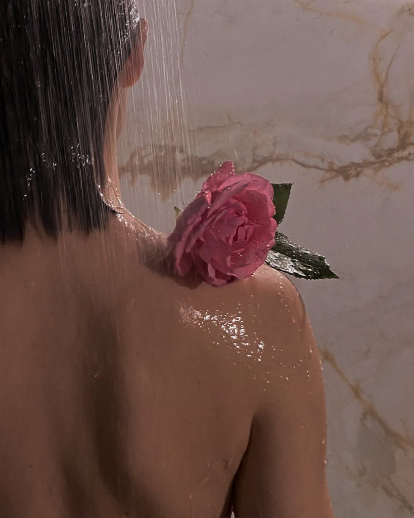 Back view of a woman showering with a pink rose on her shoulder, water cascading down.