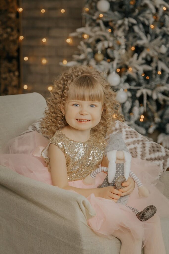 Young girl with curly blond hair and toy by a decorated Christmas tree.