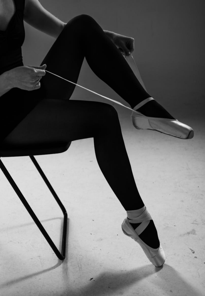 Black and white photo of a ballerina tying pointe shoes, emphasizing grace and preparation.