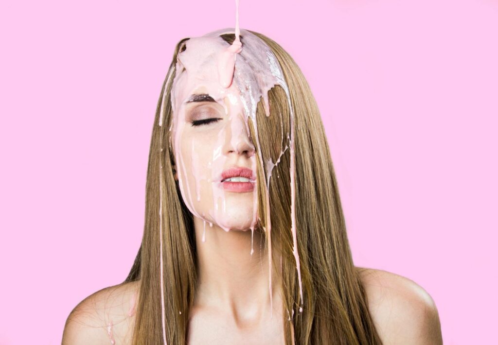 Artistic portrait of a woman with pink paint dripping on her face, against a pastel background.