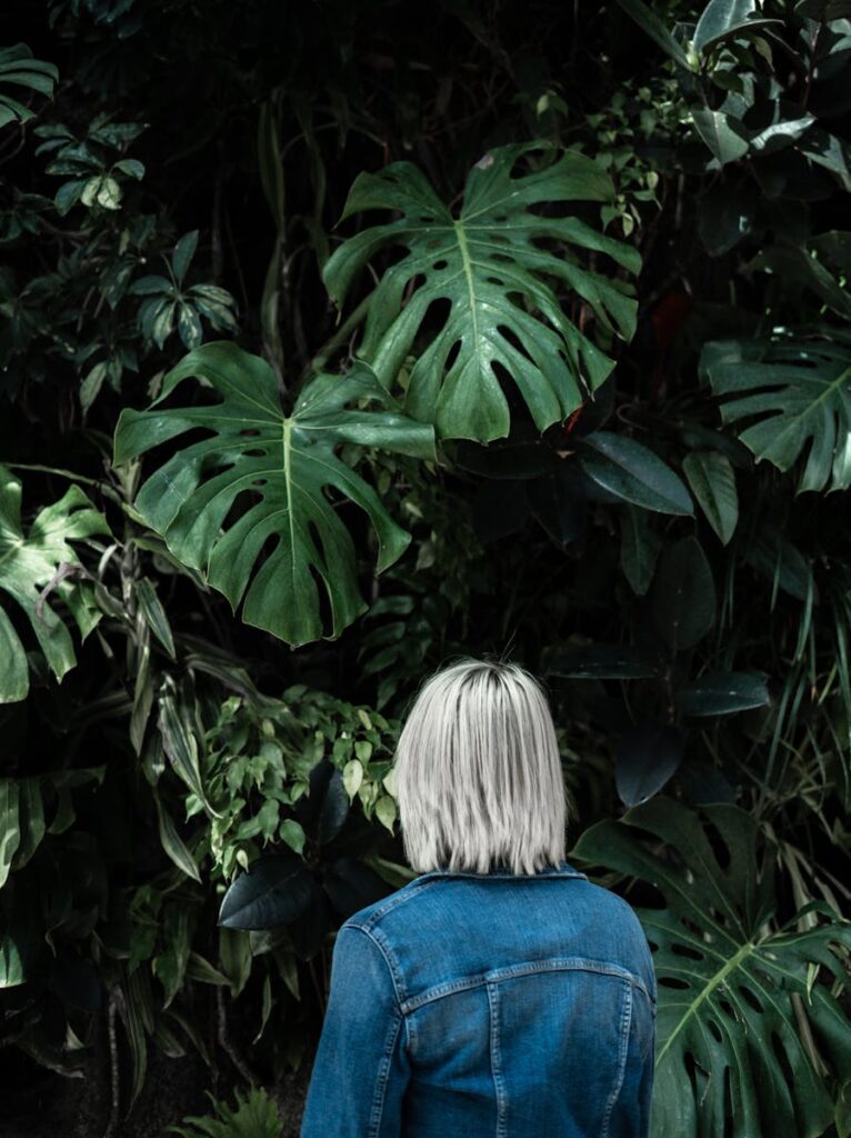 A woman in a denim jacket stands among lush tropical plants, facing away.
