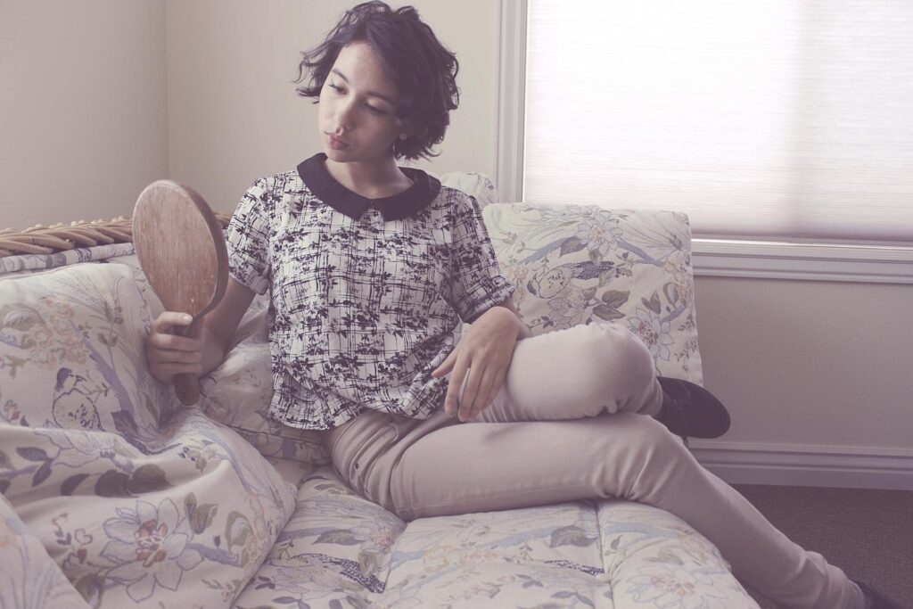 A young woman with short hair holds a mirror while sitting on a floral sofa in a cozy, light-filled room.