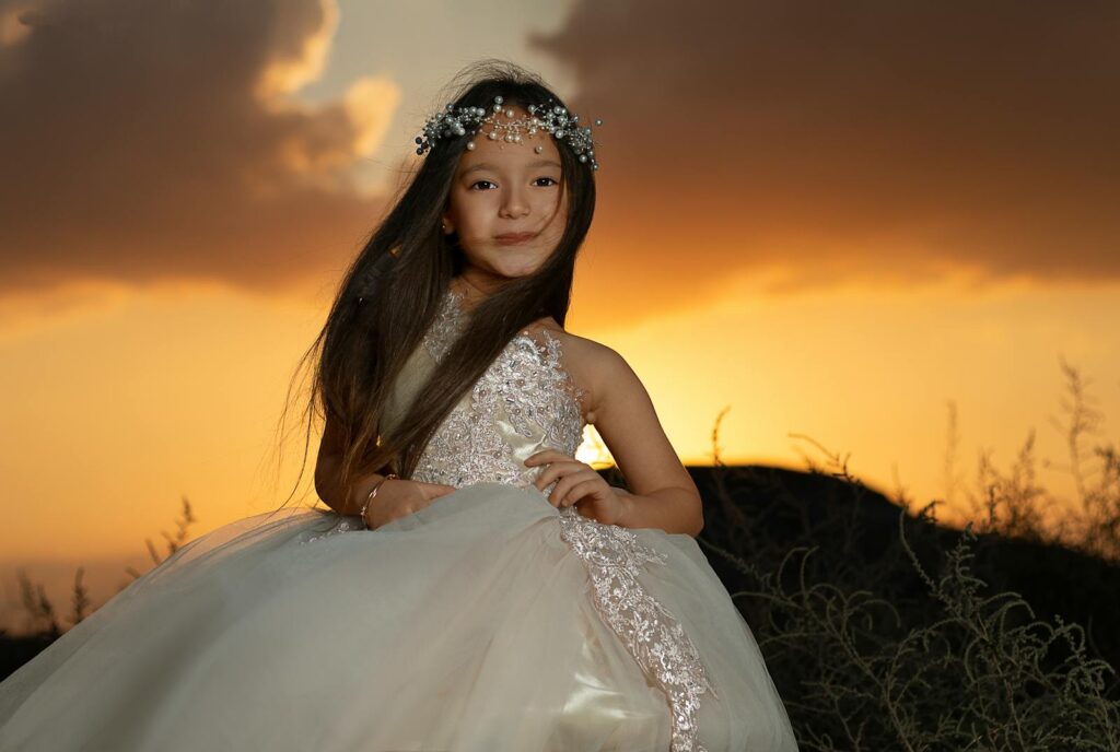 A young girl wearing a floral headpiece and white gown poses gracefully against a beautiful sunset backdrop.