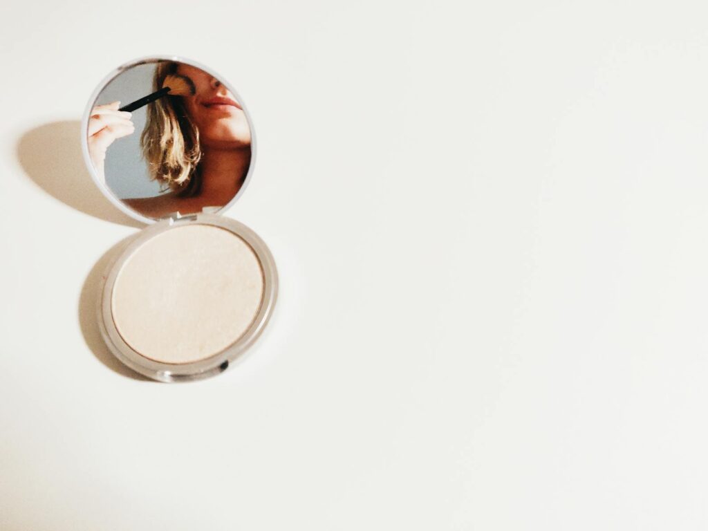 Close-up of a woman using makeup in a compact mirror, set against a bright background.