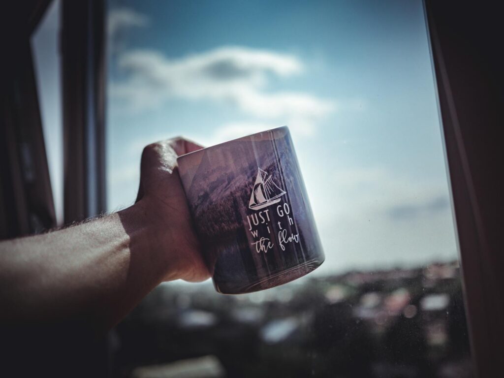 A hand holds an inspirational mug with 'Just Go With the Flow' near a window, capturing a serene view.