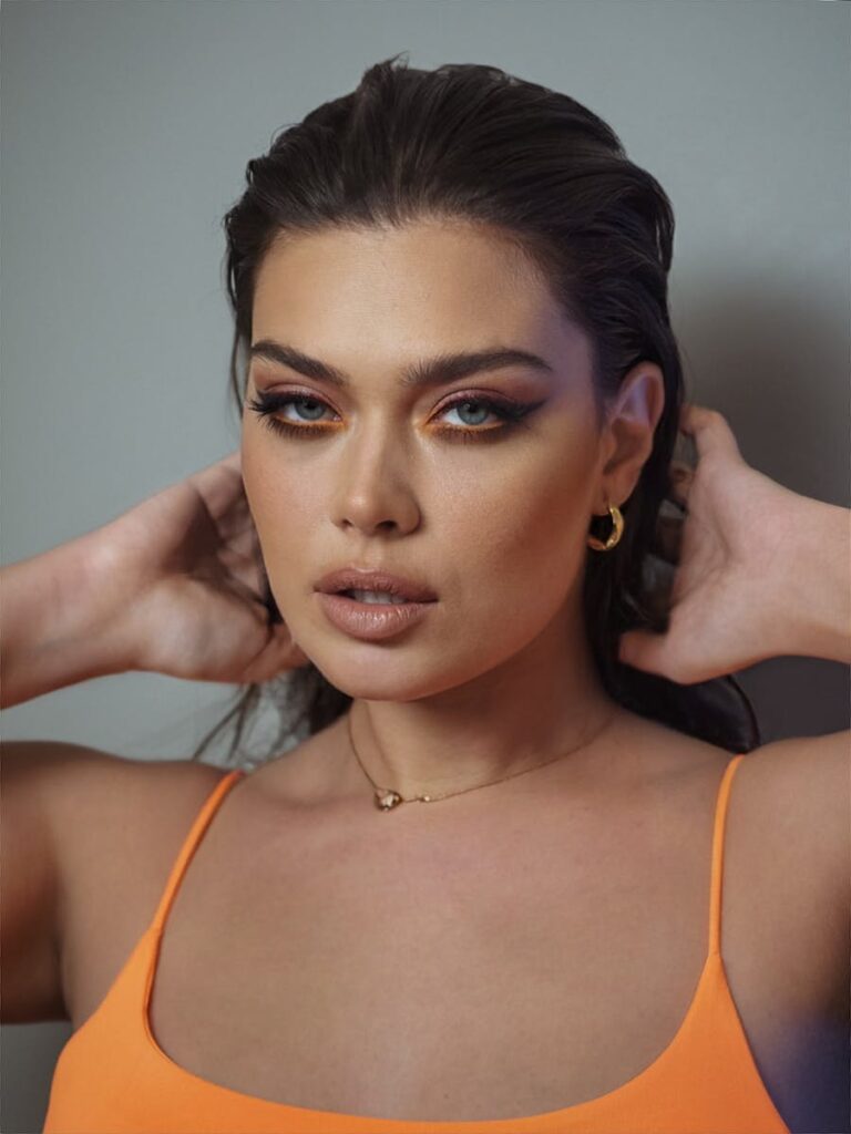 Portrait of a stylish woman in an orange outfit with bold makeup and earrings.