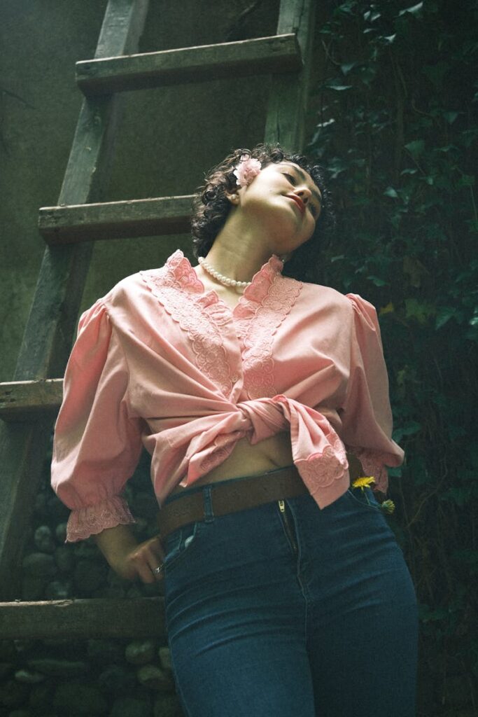 Young woman in pink blouse and jeans posing outdoors with a natural backdrop and rustic ladder.