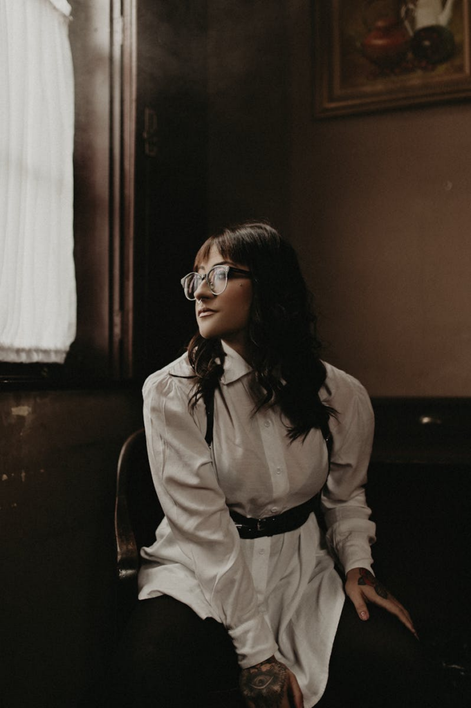 A young woman poses in a white shirt dress with a black belt, sitting near a window.