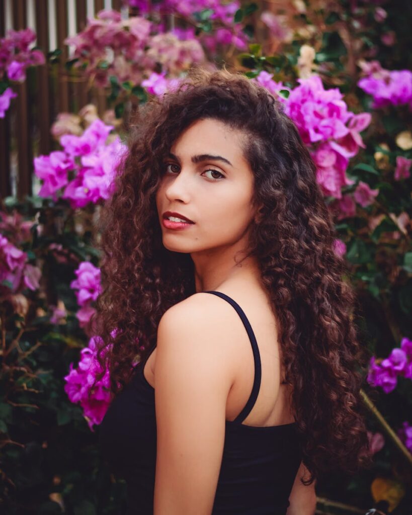 Portrait of a young woman with curly hair posing outdoors surrounded by vibrant purple flowers.