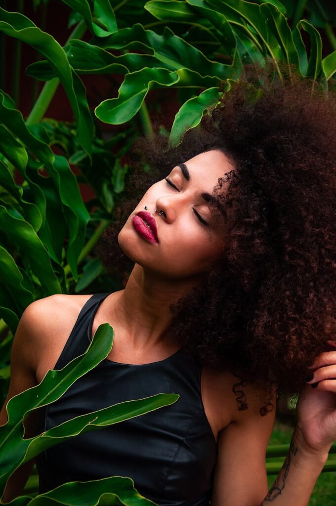 Beautiful African American woman with curly hair posing among vibrant green leaves, exuding confidence and elegance.