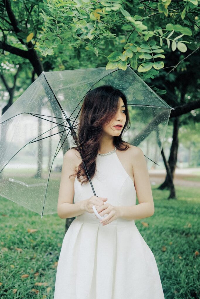 Young woman in white dress holding a transparent umbrella in a lush green park.