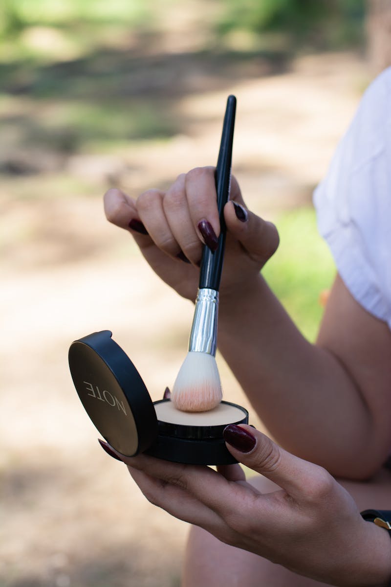 Close-up of hands applying makeup setting powder with a brush outdoors.