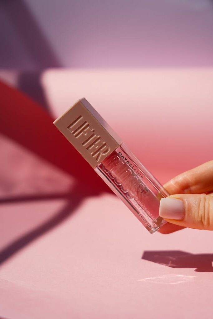 Stylish hand holding clear lip gloss against a pink backdrop.