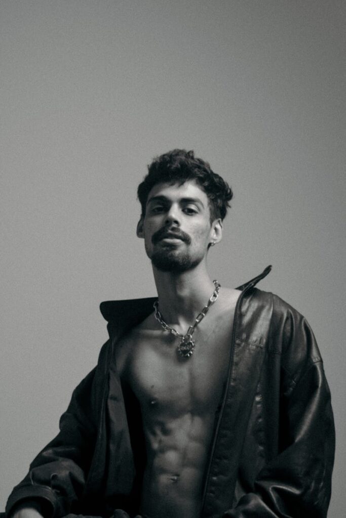 Striking black and white portrait of a shirtless man with necklace and jacket.