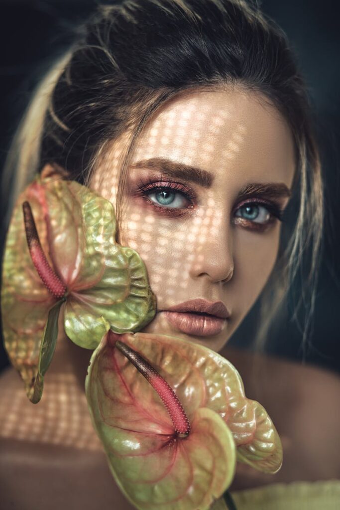 Close-up portrait of a woman with vibrant makeup and anthurium flowers, featuring dramatic lighting.