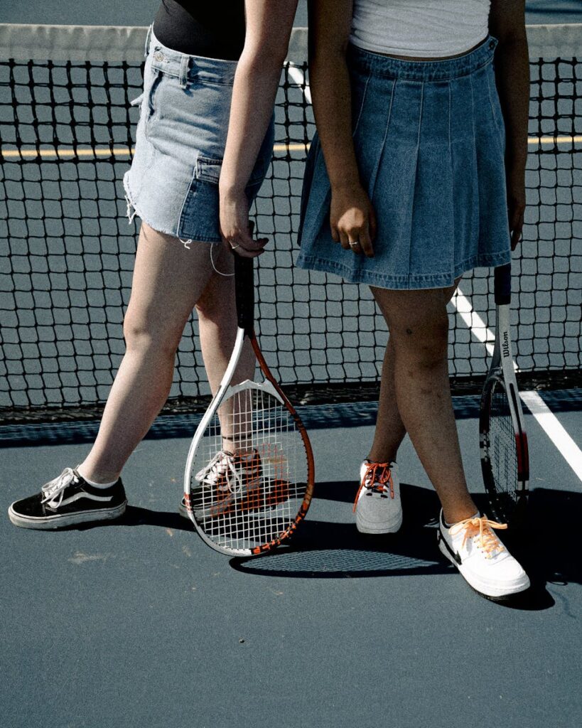 Two women in denim skirts holding tennis rackets on a sunny court.