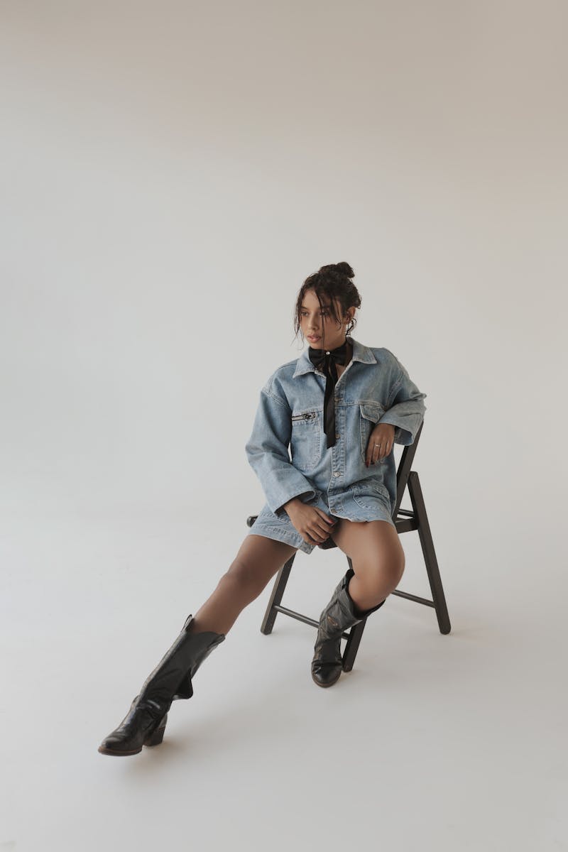 Stylish portrait of a young woman wearing a denim jacket and black boots seated on a chair.