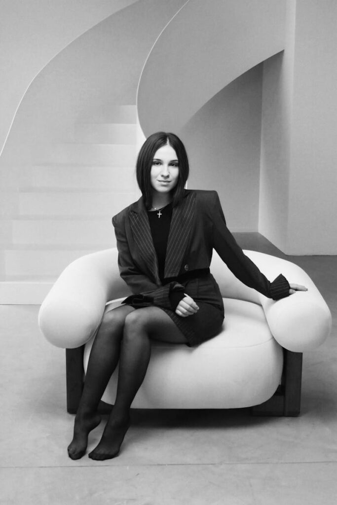 Stylish woman in a black outfit seated in a modern room with spiral staircase.