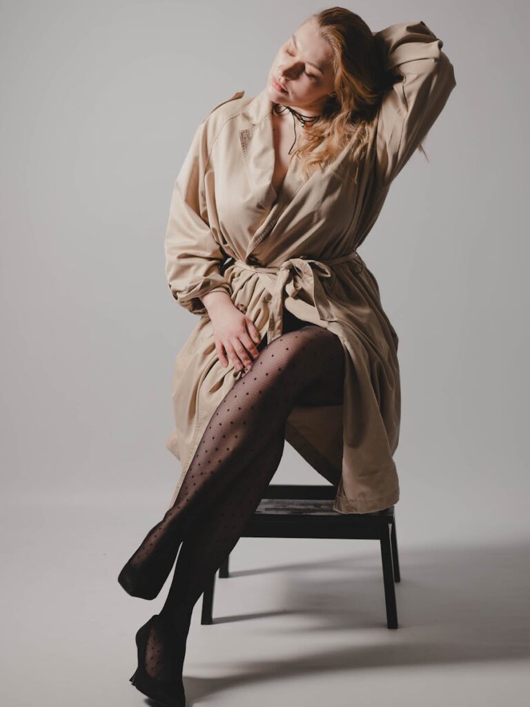 Elegant woman in a beige trench coat with black tights, sitting gracefully in studio.