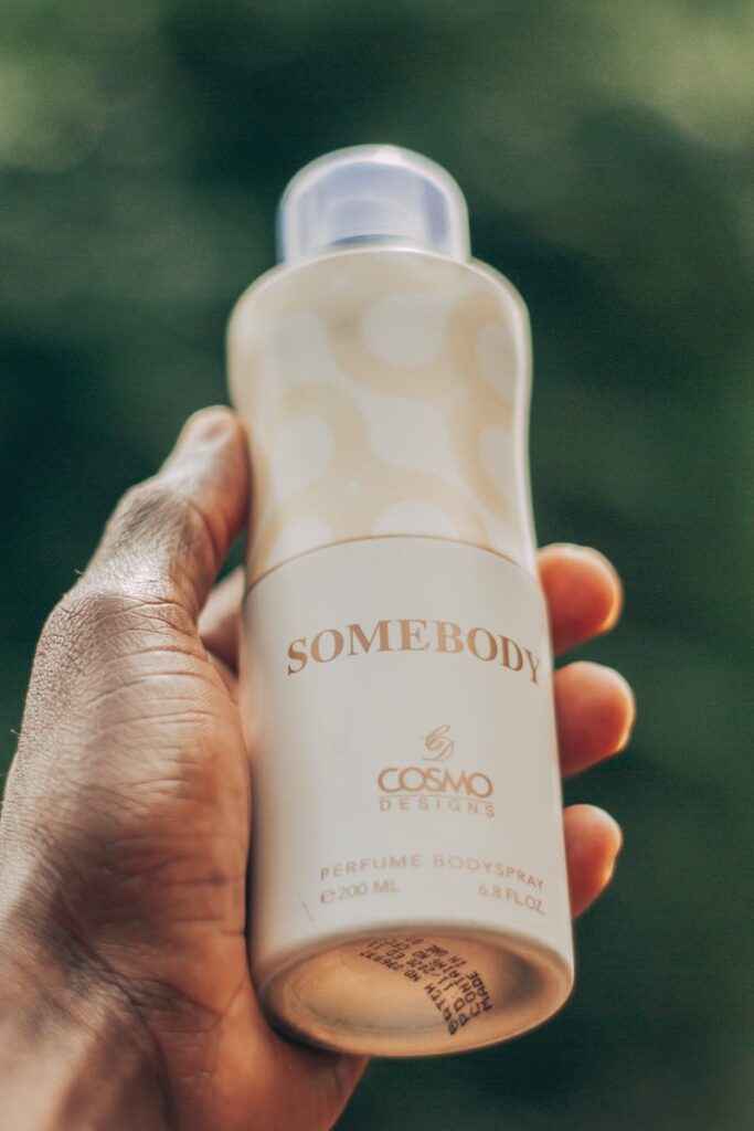 A close-up of a hand holding Somebody perfume body spray with a blurred green background.