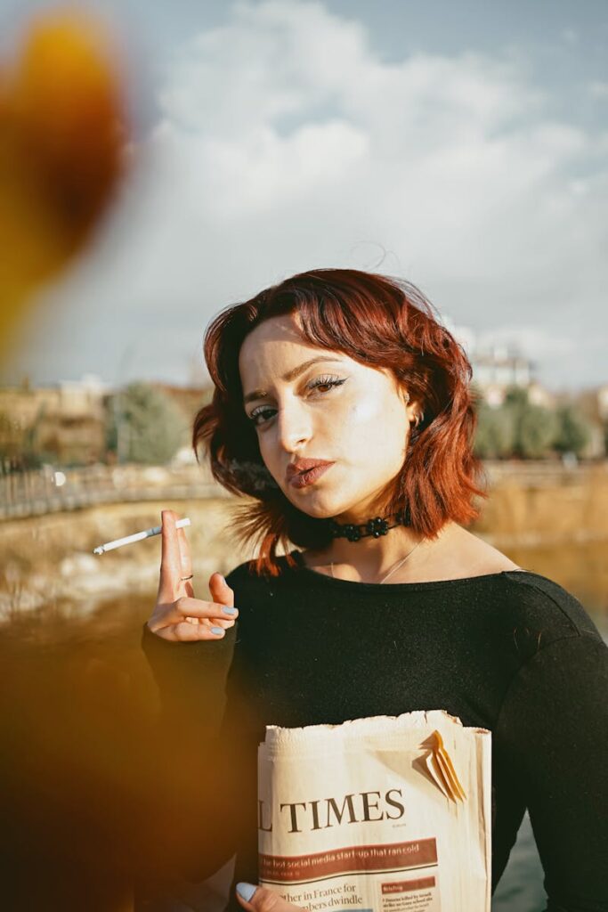 A woman with red hair holding a newspaper and cigarette outdoors on a sunny day.