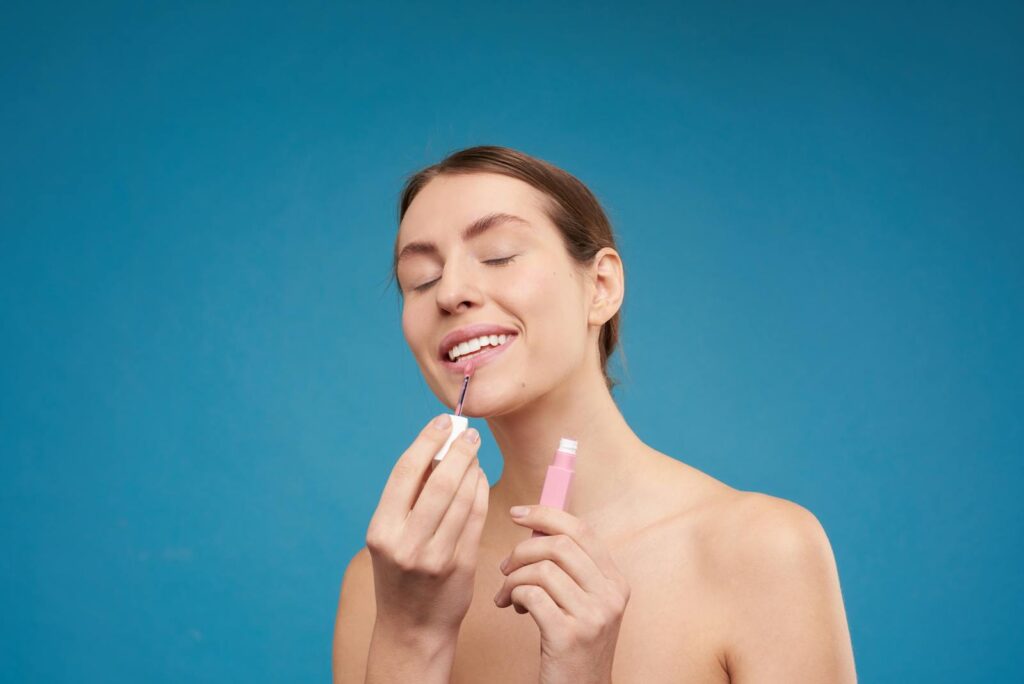 A smiling woman applying lip gloss with closed eyes on a blue background.