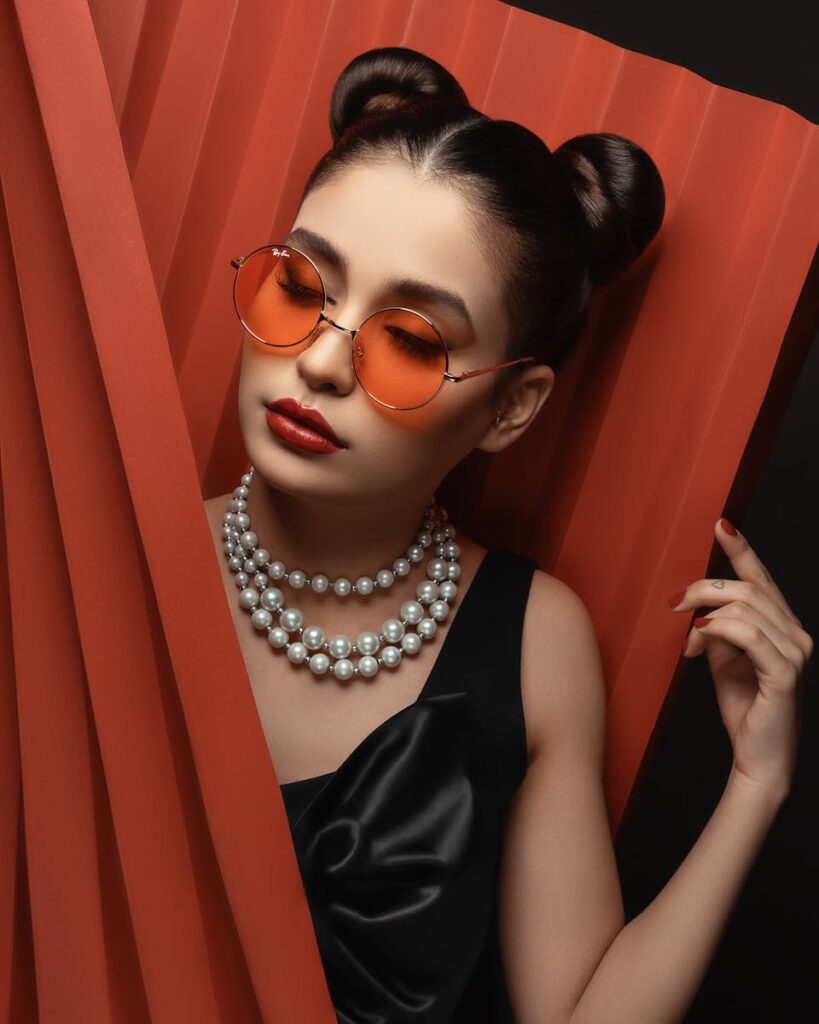 Stylish woman in black dress, pearls, and orange sunglasses posing gracefully.