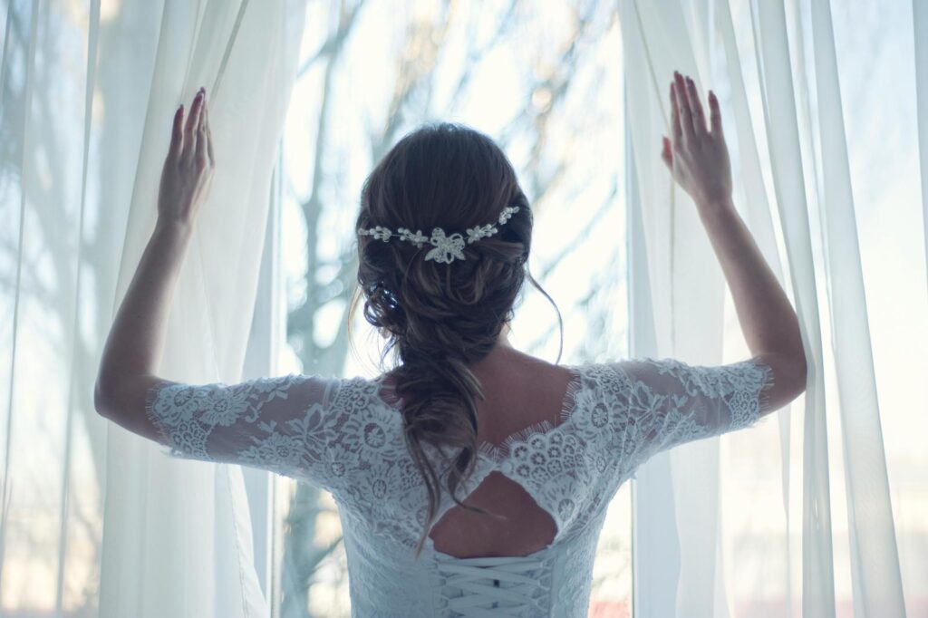 Beautiful bride in lace dress looks out a sunlit window, capturing a moment of serene elegance.