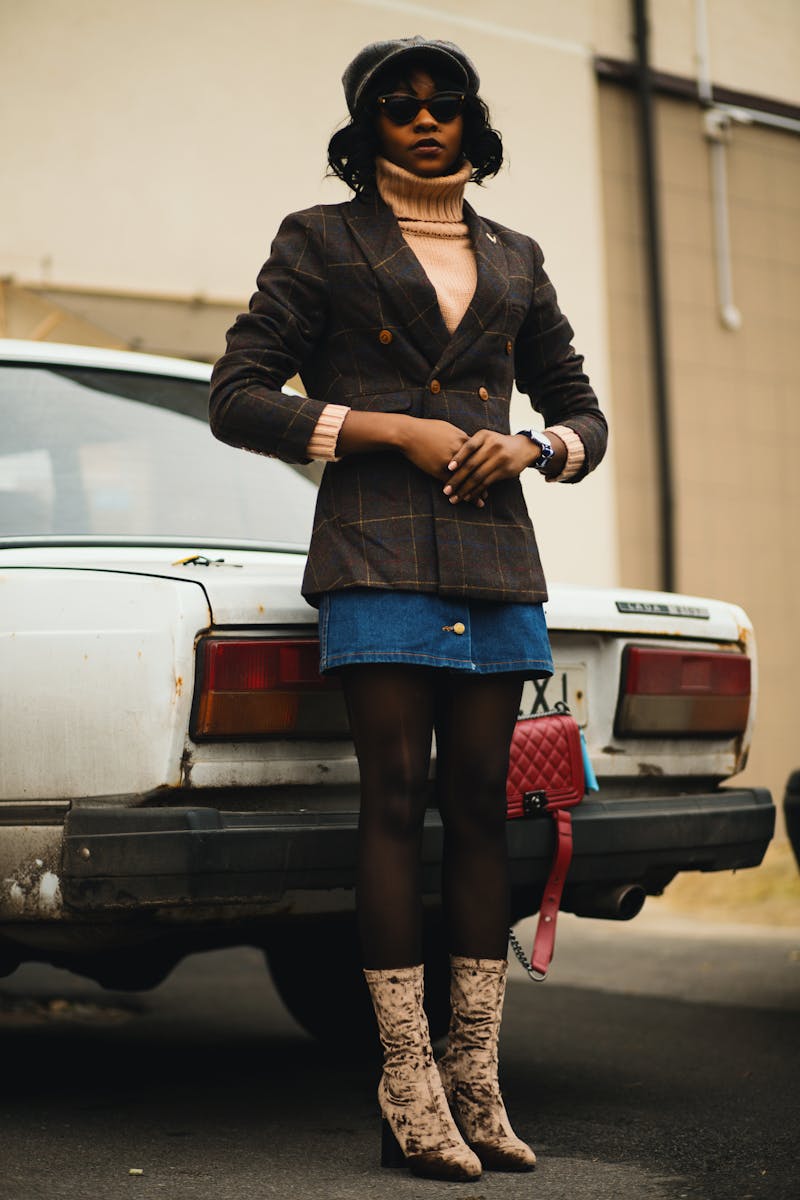 Stylish black woman in turtleneck and blazer poses next to a car showcasing vintage fashion.
