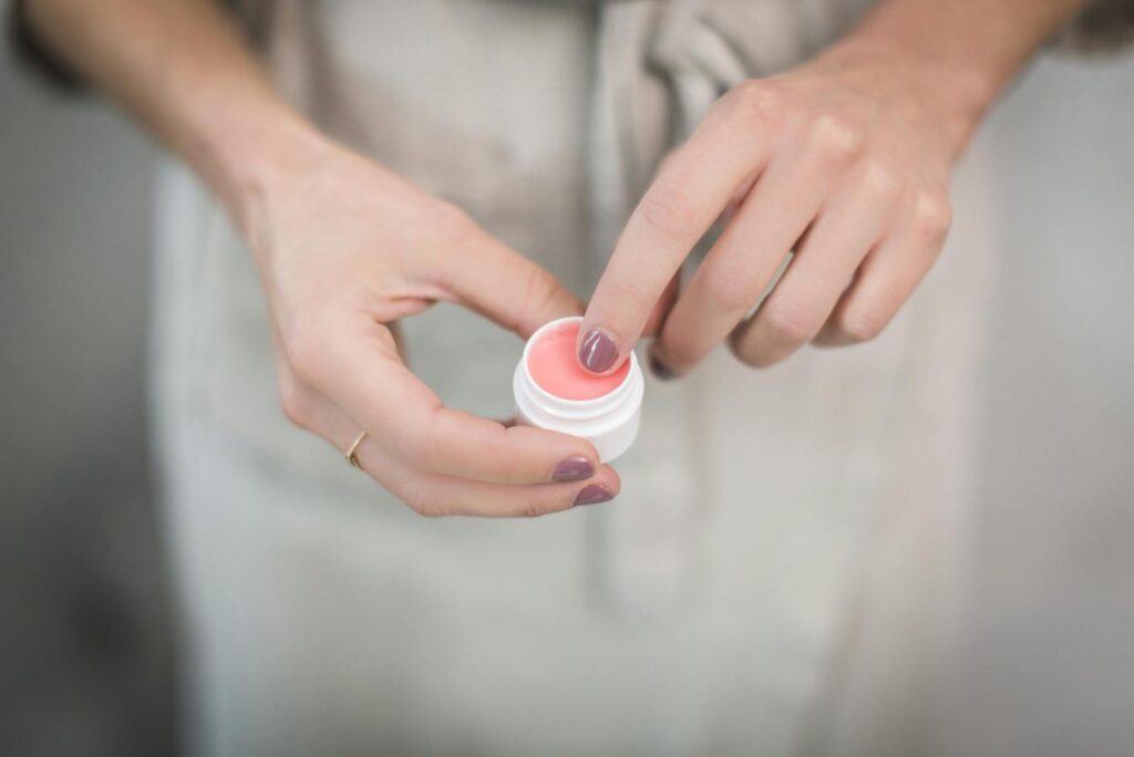 Close-up of woman's hands applying pink lip balm from a small jar, highlighting skincare routine.