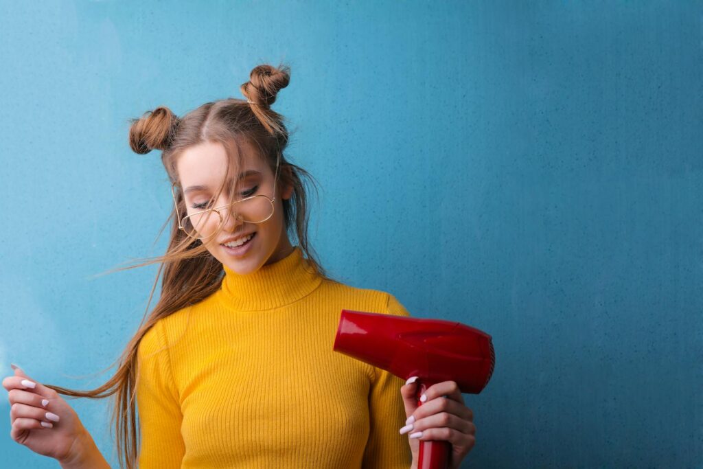 Woman with quirky hairstyle and glasses smiling with a hair dryer against a blue background.