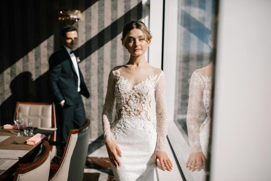 Romantic indoor wedding photoshoot displaying the bride in an elegant gown with groom in background.