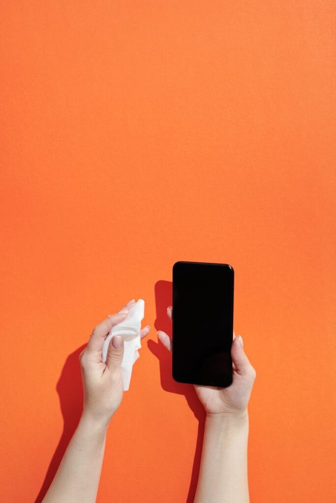 Hands cleaning a smartphone with wipes against a vibrant orange background, emphasizing hygiene.