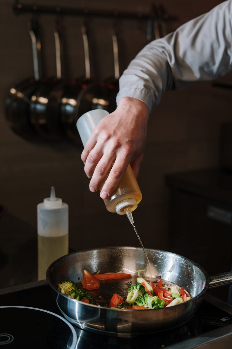 A chef's hand drizzling oil over mixed vegetables in a frying pan in a kitchen setting.