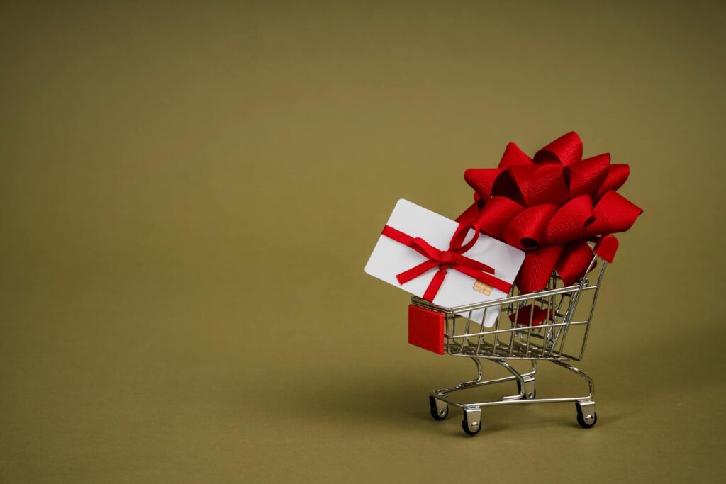 Mini shopping cart containing a gift card adorned with a red bow on a neutral background, ideal for retail concepts.