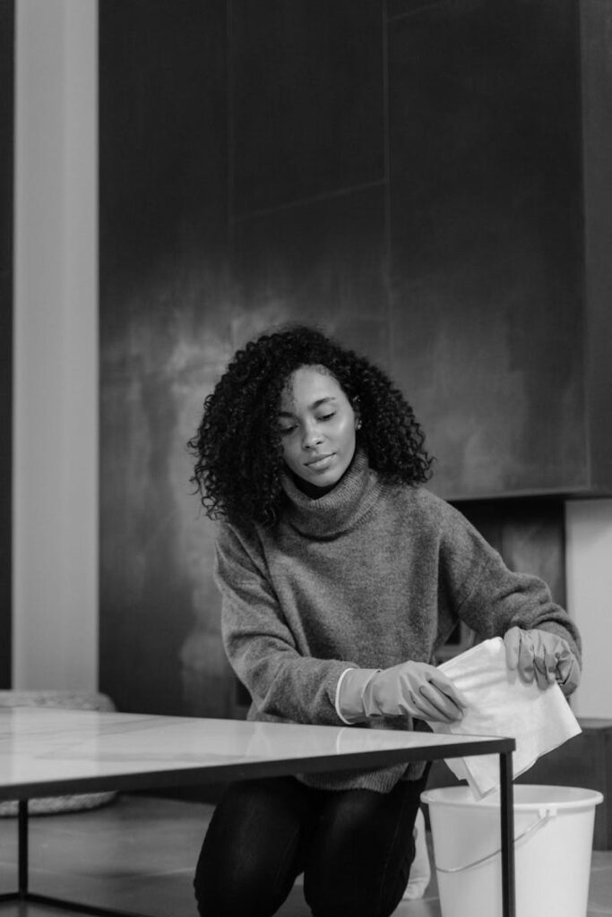Black and white photo of a woman with curly hair cleaning a table indoors.