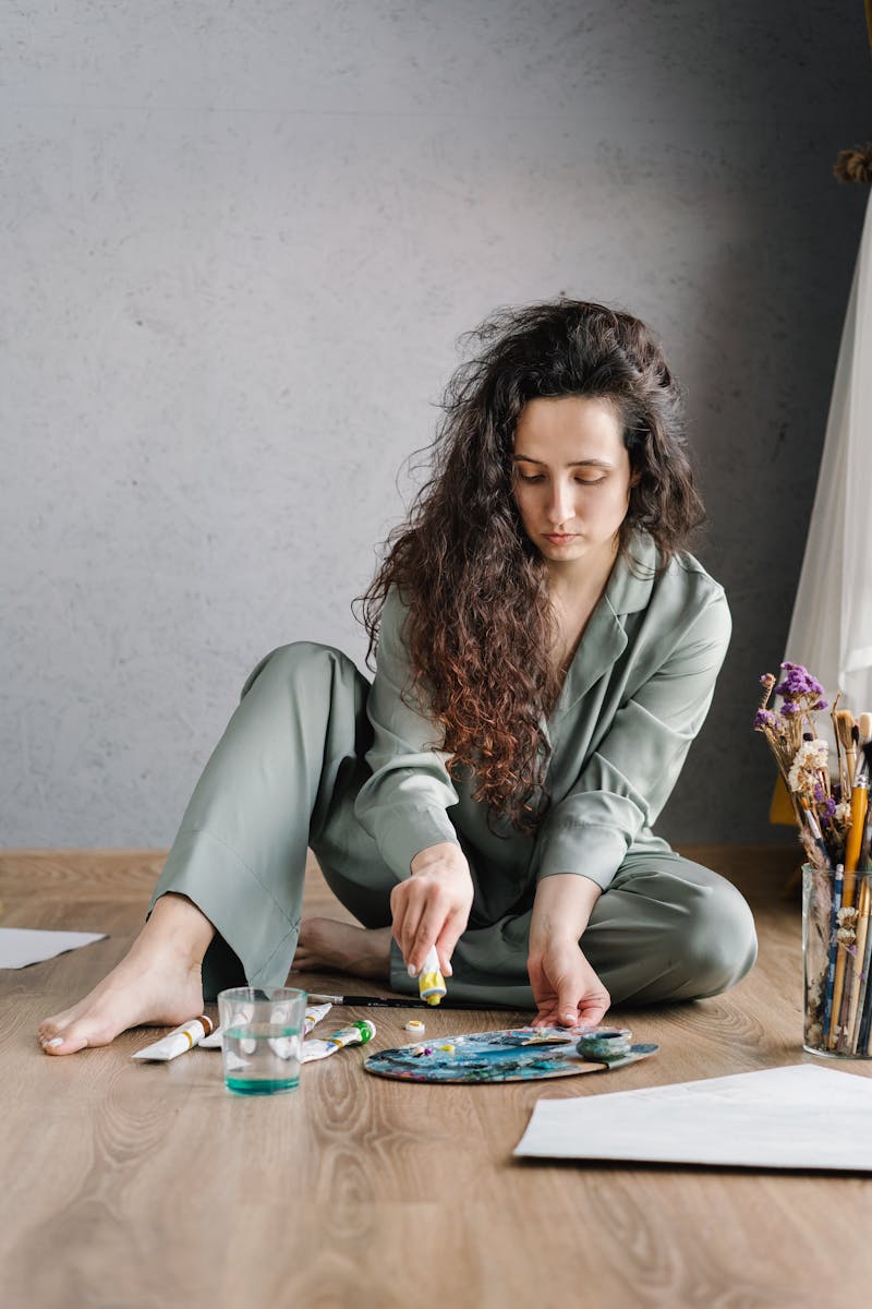 A young woman artist in pajamas working on her painting on a wooden floor indoors.