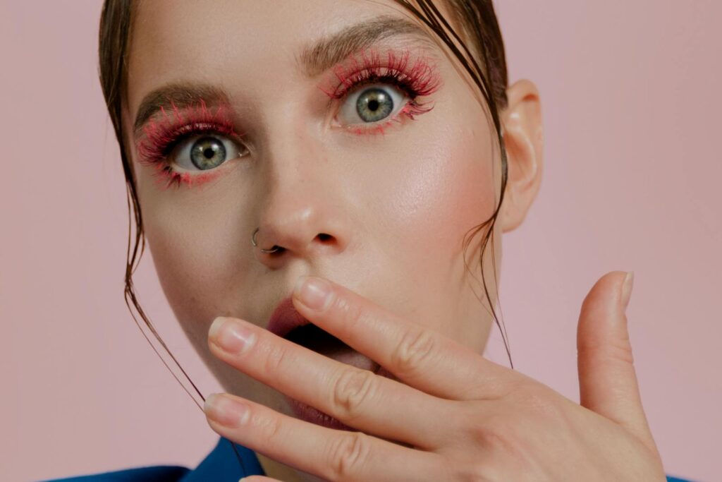 Close-up portrait of an adult with dramatic pink eye makeup on a pink background, expressing surprise.