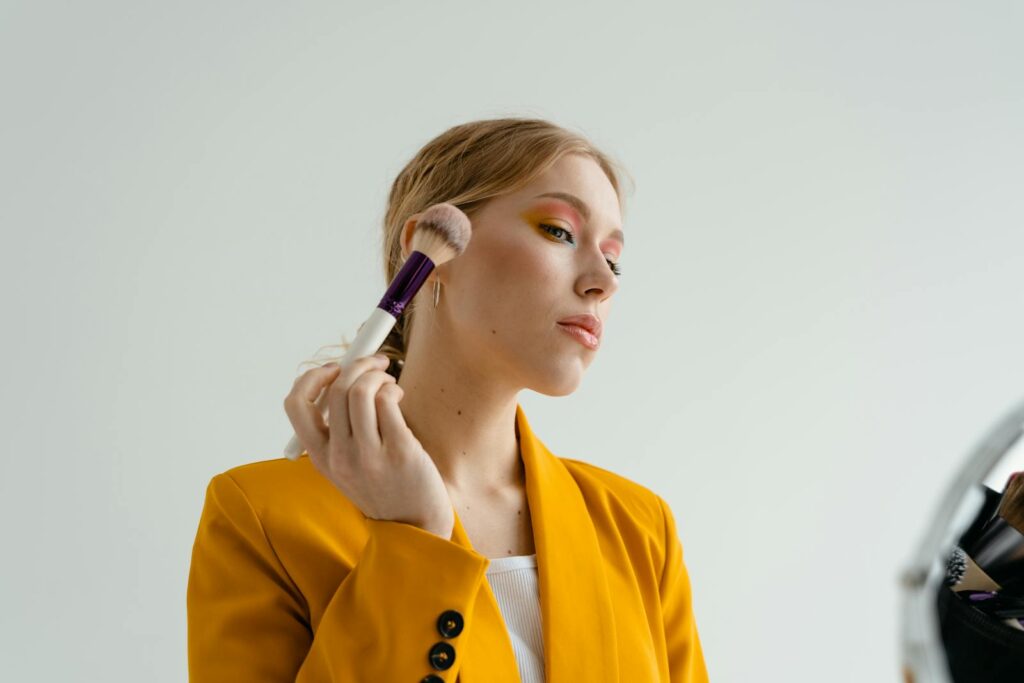 A woman in a yellow blazer applies makeup with a brush, showcasing a glamorous look.