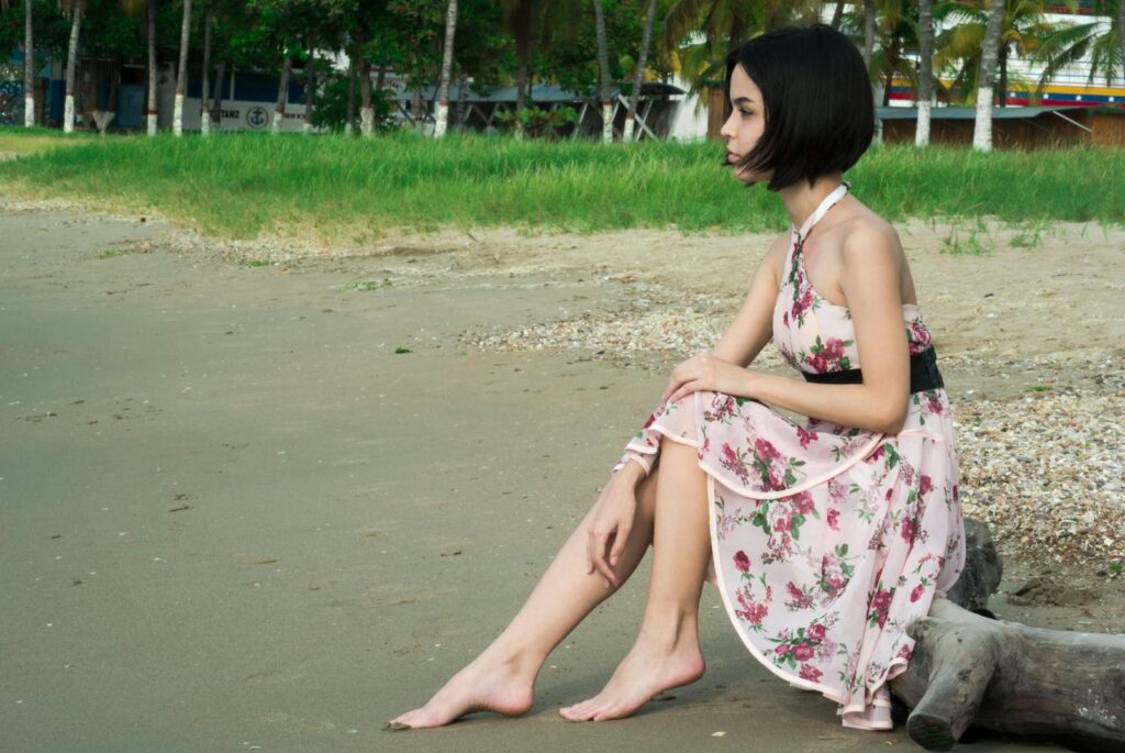 A young woman with short hair sits barefoot on a sandy beach wearing a floral dress.