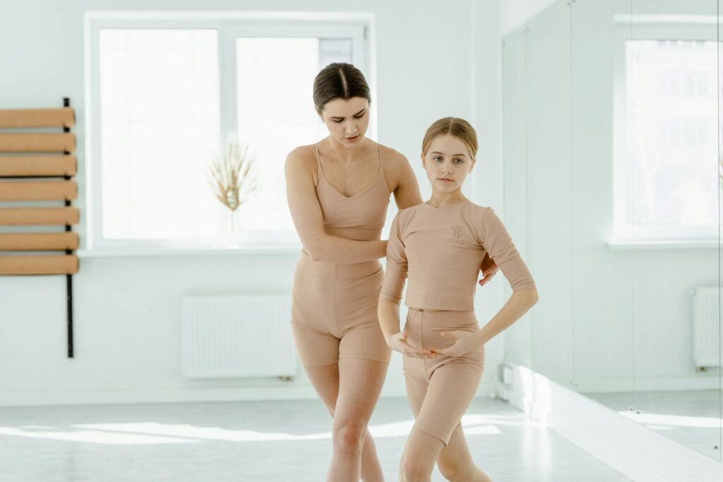 A ballet teacher guides a young girl in a bright dance studio, emphasizing technique and elegance.