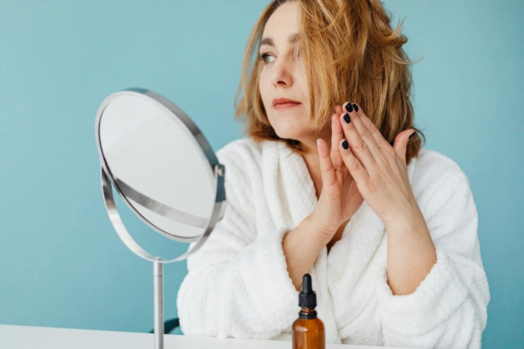 Woman in white robe applying skincare oil, looking in the mirror with manicured nails.