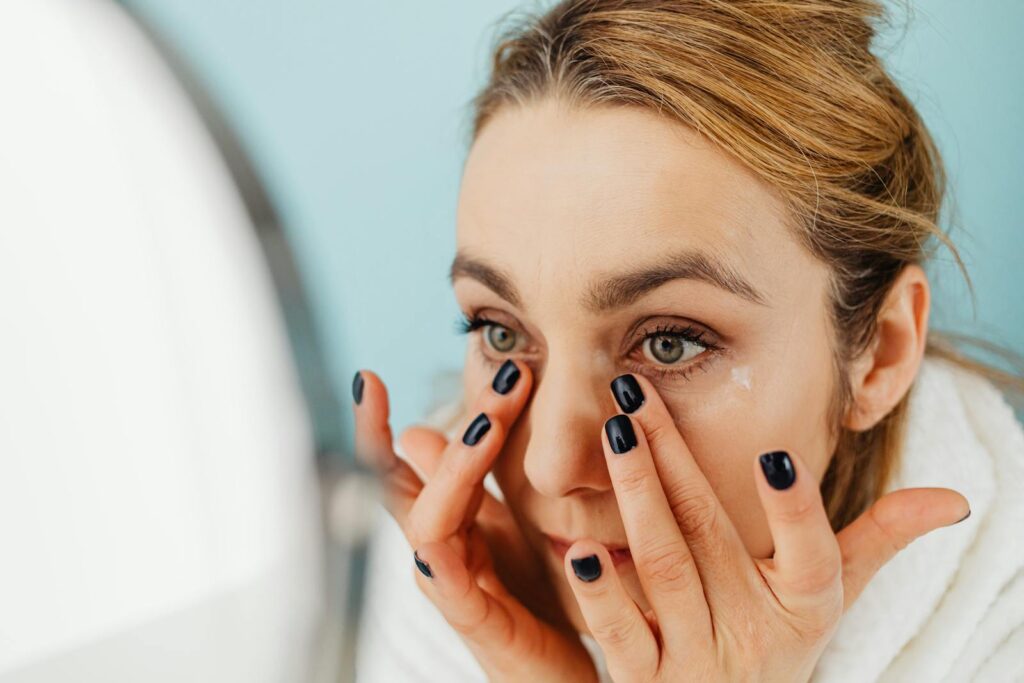 Close-up of a woman applying facial cream in front of a mirror with blue background.