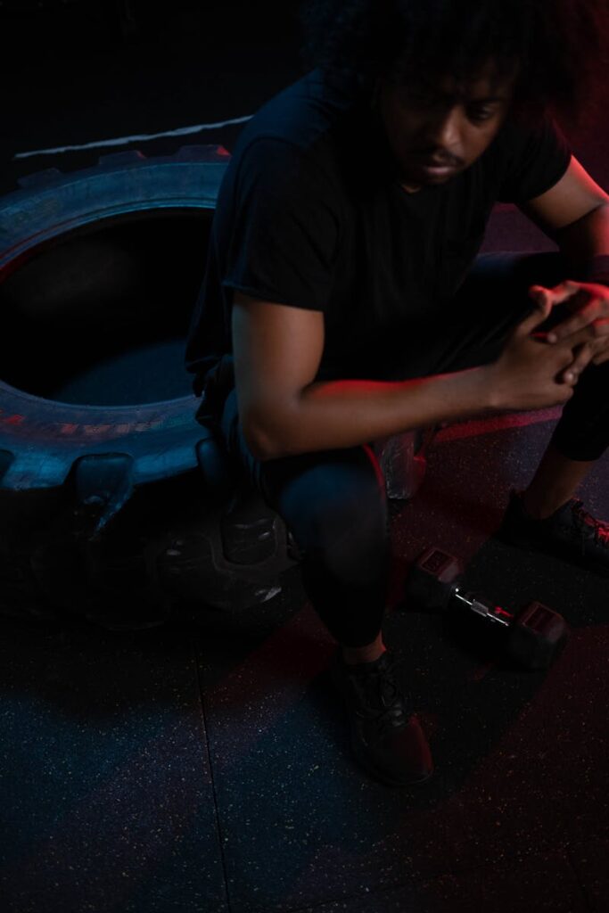An athlete with afro hair rests on a tire in a dimly lit gym, clutching hands thoughtfully.