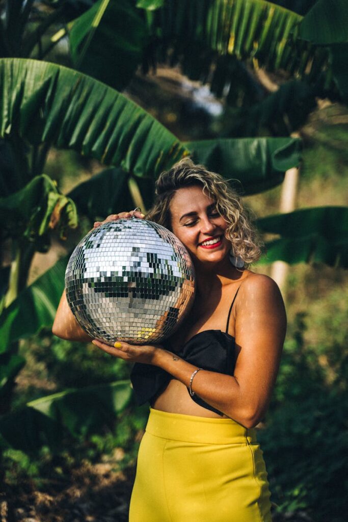 A happy woman with blonde curls holds a disco ball amidst lush green foliage, embracing a vibrant outdoor setting.