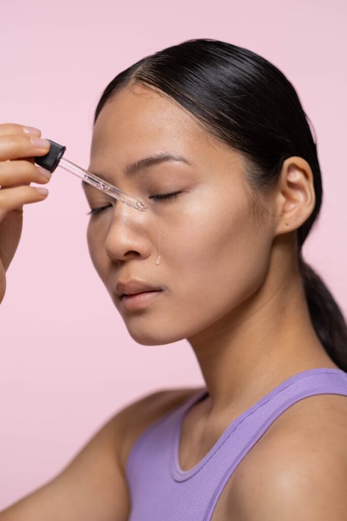 Close-up of a woman applying facial serum using a dropper on a pink background.