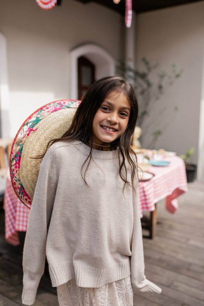 A cheerful girl wearing a straw hat and sweater smiles indoors by a table.