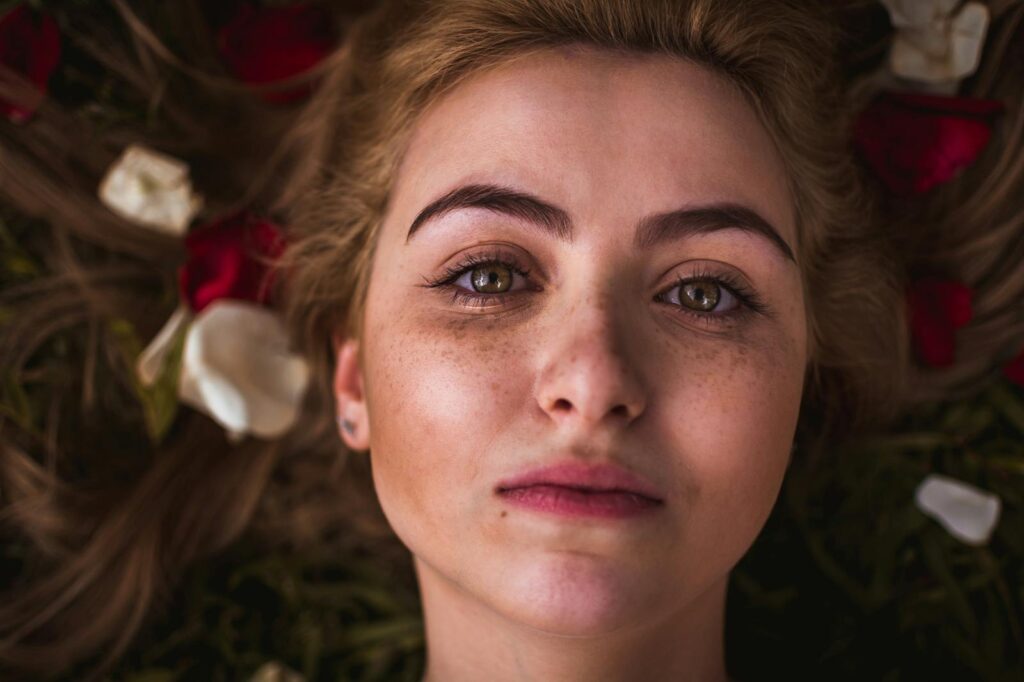 A striking close-up portrait of a freckled woman surrounded by rose petals.