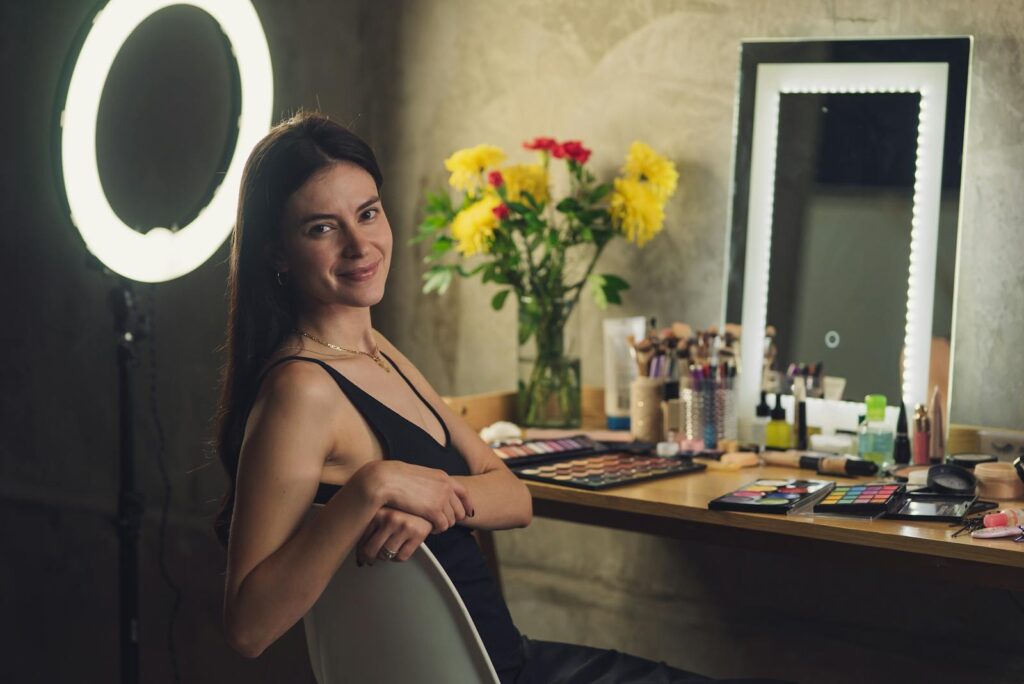 Woman sitting at a makeup station with flowers, cosmetics, and a ring light, smiling at the camera.