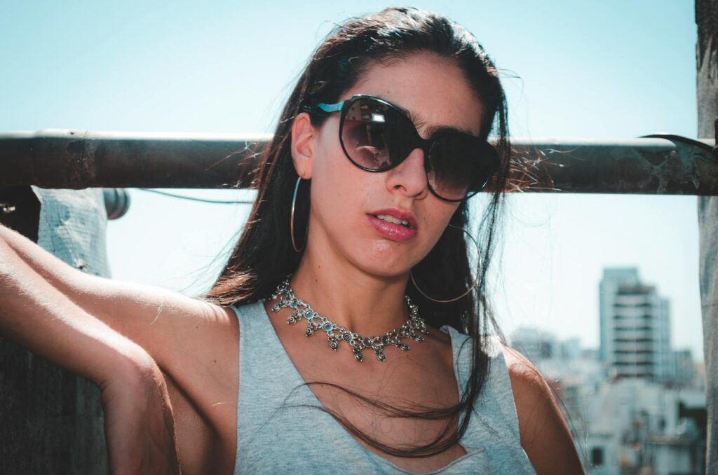 Elegant woman posing outdoors wearing sunglasses and a necklace against a city skyline.